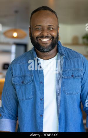Uomo afroamericano che sorride mentre è in piedi in una cucina aperta moderna, indossa una camicia di jeans blu. Ritratto, stile di vita, calore, interno, ambiente, piano Foto Stock