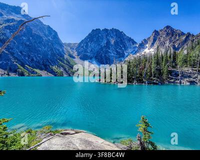 Lago turchese riflette montagne frastagliate sotto un cielo blu terso. Sempreverdi rigogliosi incorniciano il bordo dell'acqua con grazia naturale. Le rive rocciose invitano a un tranquillo c Foto Stock