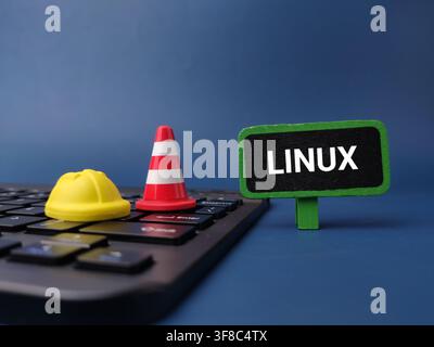 Un casco di sicurezza in miniatura giallo, un cono di segnalazione stradale e un cartello con la scritta LINUX poggiano su una tastiera del computer scura. Foto Stock