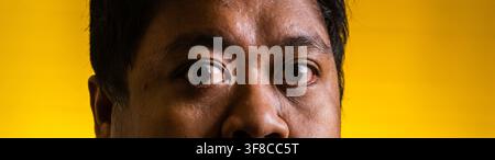 Ritratto macro che dettagli il intenso sguardo di un uomo asiatico adulto su uno sfondo giallo brillante. La composizione strettamente ritagliata si concentra rigorosamente Foto Stock