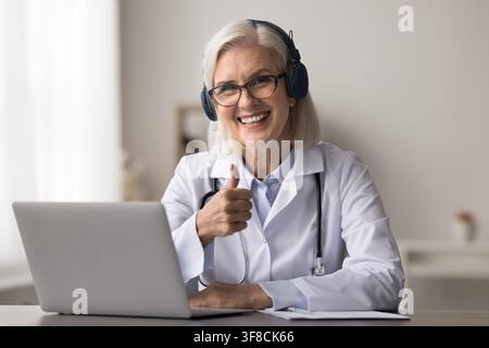 Medica anziana indossa cuffie e mostra il pollice alzato sul posto di lavoro Foto Stock