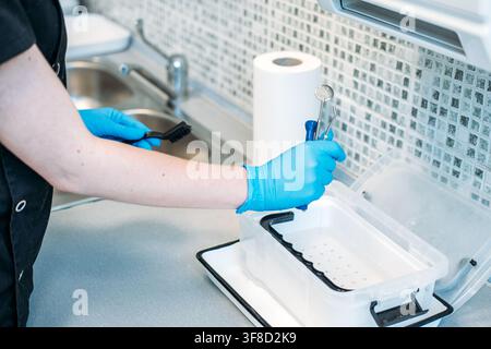 Persona che indossa guanti protettivi solleva strumenti metallici da un contenitore di plastica tenendo una spazzola nell'altra mano. Fase di pulizia pre-sterilizzazione, dentale Foto Stock