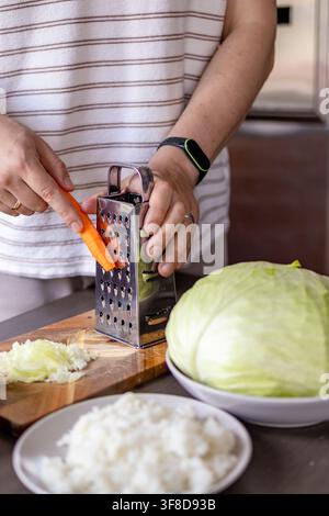 Uomo gratta le carote accanto al cavolo e ad altri ingredienti mentre prepara involtini di cavolo fatti in casa. Concetto di lavoro condiviso in cucina, preparazione del pasto, fam Foto Stock