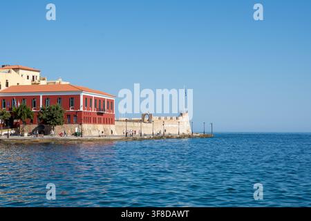 Vista dell'edificio storico rosso e delle fortificazioni in pietra al porto della vecchia città di Chania con persone che passeggiano lungo il lungomare a Chania, Creta, Grecia Foto Stock