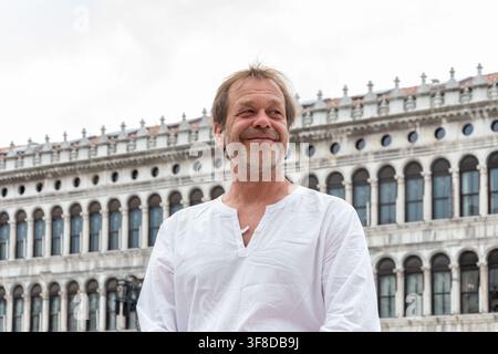 Un uomo maturo sorridente con una camicia bianca si trova all'aperto davanti a un edificio ad arco storico sotto un cielo nuvoloso. Foto Stock