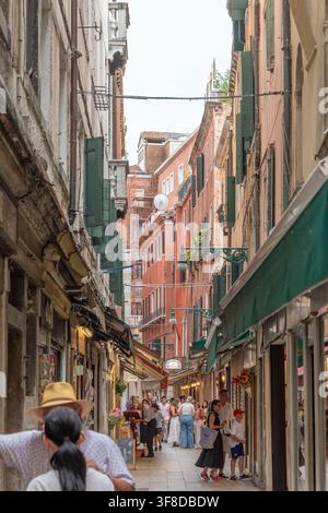 VENEZIA, ITALIA, 11 luglio 2025: Persone camminano lungo una stretta vecchia contrada tra alti edifici con tende da negozio e persiane verdi. Foto Stock