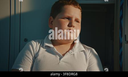 Primo piano di un bambino triste seduto sul pavimento del corridoio vicino a un armadietto aperto con espressione pensierosa durante la ricreazione. Benessere emotivo, routine scolastica, sfide dell'infanzia, benessere mentale, educazione. Foto Stock