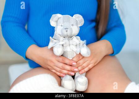 Una madre in attesa in maglietta blu tiene delicatamente un orsacchiotto a quadri contro il ventre. Foto Stock