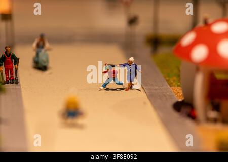 Una foto ravvicinata mostra figure in miniatura dipinte su una strada modello, con una donna anziana che si protende verso un bambino che corre vicino al marciapiede. Foto Stock