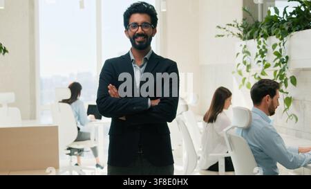 Uomo d'affari arabo-indiano CEO sicuro di sé che sorride guardando la telecamera in ufficio, capo squadra, datore di lavoro, dirigente esecutivo, in posa per un ritratto aziendale con le braccia Foto Stock