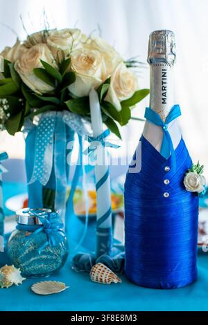 Una bottiglia di champagne blu decorata è posizionata accanto a un bouquet nuziale, una candela decorativa e conchiglie marine. Foto Stock
