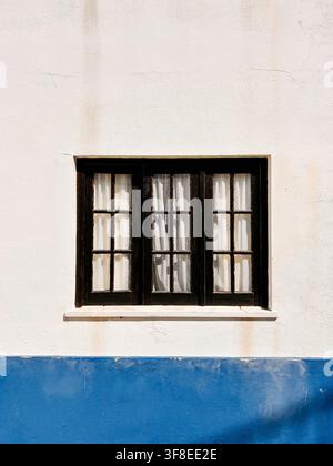 Una finestra tradizionale incorniciata di nero con tende bianche inserita in una semplice facciata bianca con una sezione inferiore dipinta di un blu vivace Foto Stock