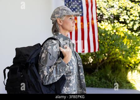 Donna adulta in mimetica digitale che porta uno zaino tattico nero su un portico vicino alla bandiera americana Foto Stock