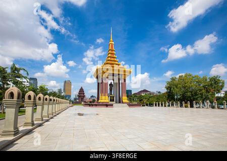 Monumento del padiglione dorato con statua in piedi nella piazza di Phnom Penh sotto un cielo blu limpido e lo skyline della città, alberi tropicali e dettagli ornati del tetto Foto Stock