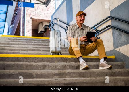Un uomo anziano è seduto alla stazione ferroviaria e aspetta di salire a bordo. Sta utilizzando un tablet digitale. Le persone anziane viaggiano. Foto Stock