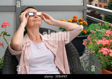 Donna di mezza età seduta su una sedia sul balcone, indossa occhiali da eclissi solare, sorride e guarda il cielo circondata da piante in vaso Foto Stock