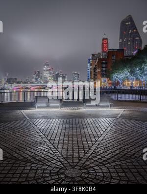 Vista dell'Oxo Tower e della skyline illuminata di Londra attraverso il Tamigi di notte, con panchine vuote su un marciapiede pavimentato lungo il fiume a Lon Foto Stock
