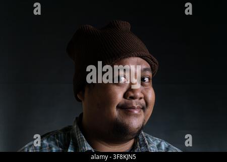 Un ritratto amichevole di un giovane uomo del sud-est asiatico dell'Indonesia che indossa un berretto di lana marrone arrotolato e una camicia a quadri casual. La persona offre Foto Stock