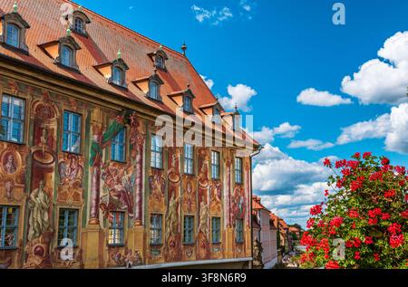 Vista grandangolare del municipio storico di Bamberg con la sua facciata affrescata riccamente dipinta e il tetto di tegole rosse. Fiori rosso acceso incorniciano il lato destro Foto Stock
