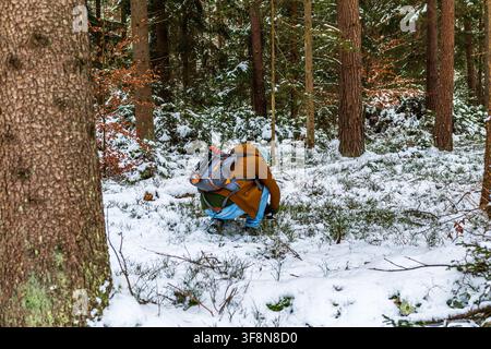 Un escursionista adulto che indossa una giacca marrone e pantaloni blu si accovaccia sul terreno coperto di neve in una foresta mista di conifere in Germania. La persona porta una Gr Foto Stock