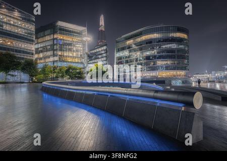 Londra, Regno Unito - 21 settembre 2024: Vista dello Shard che si erge sopra moderni edifici di uffici in vetro e una passerella curva illuminata di notte. Foto Stock