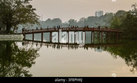 Hanoi, Vietnam - 16 gennaio 2025: Vista del ponte Huc con persone che attraversano la struttura in legno rosso sopra il lago Hoan Kiem, circondato da alberi. Foto Stock