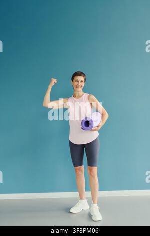 Ritratto di una donna caucasica anziana sorridente che tiene un tappetino da yoga e flette il bicipite contro un muro blu, promuovendo l'invecchiamento attivo e i programmi di fitness per anziani, verticale Foto Stock