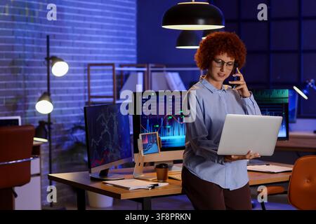 Analista finanziaria con capelli rossi ricci che lavora fino a tardi in un ufficio buio, tiene un laptop e parla al telefono cellulare Foto Stock