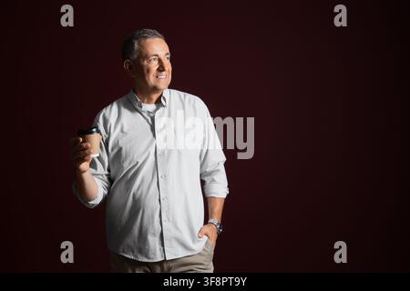 Uomo maturo sorridente che tiene una tazza da caffè monouso, guarda lontano con un'espressione pensierosa, su uno sfondo scuro Foto Stock