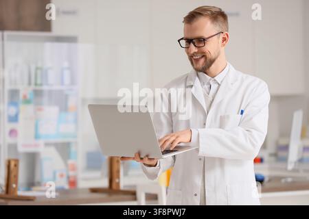 Farmacista maschio sorridente in camice bianco e occhiali che utilizza un laptop in un ambiente moderno di farmacia, È impegnato in compiti digitali, l'integrazione di te Foto Stock