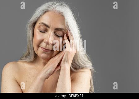 Donna anziana con capelli grigi che si prende cura della pelle con crema per il viso, concetto di spa domestica Foto Stock