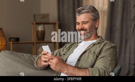 Uomo caucasico di mezza età sorridente che usa uno smartphone sul divano a casa, concetto di stile di vita rilassato Foto Stock