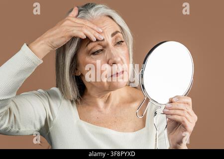 Donna anziana caucasica che ispeziona le rughe allo specchio a casa, concetto di routine di cura della pelle Foto Stock
