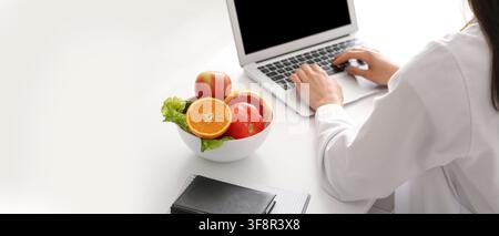 Nutrizionista o dietista che lavora su un laptop con una ciotola di frutta e verdura fresca su una scrivania bianca, alimentazione sana e benessere Foto Stock