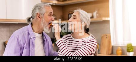 Coppia anziana felice che si gode uno spuntino insieme in cucina, sorridendo e nutrendosi a vicenda Foto Stock