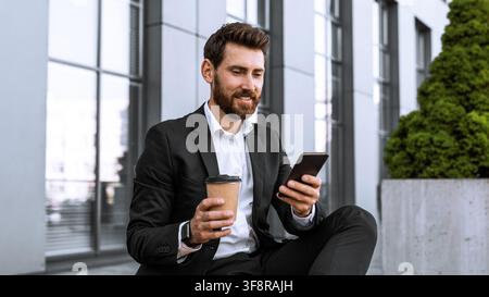 Uomo barbuto sorridente in abito che usa uno smartphone mentre beve caffè, concetto di ufficio moderno Foto Stock