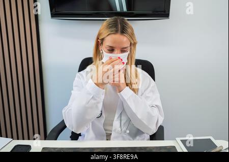 Dottora che regola una mascherina al tavolo in uno studio medico, provando stress e stanchezza Foto Stock