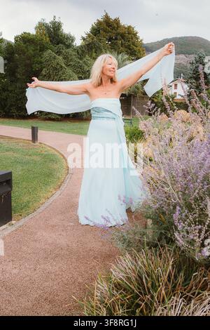 Donna in un vestito blu fluente che posa gioiosamente con le braccia alzate, abbracciando la natura in una bella ambientazione di giardino. La sua espressione riflette felicità e f Foto Stock