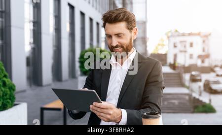 Uomo caucasico felice che utilizza un tablet fuori dall'edificio dell'ufficio, concetto aziendale Foto Stock