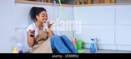Donna afroamericana sorridente che pulisce la cucina mentre parla al telefono, concetto di pulizia domestica Foto Stock
