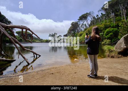 Donna in piedi vicino a un lago, che filma il paesaggio naturale con un telefono cellulare Foto Stock