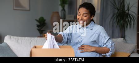 Donna afroamericana felice che disimpacchetta vestiti da bambino a casa, concetto di maternità in attesa Foto Stock