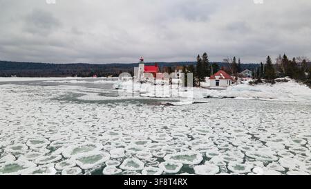 Vista aerea del faro storico di Eagle Harbor circondato da banchi di ghiaccio sul lago ghiacciato sotto un cielo invernale nuvoloso a Eagle Harbor, Michigan, Un Foto Stock