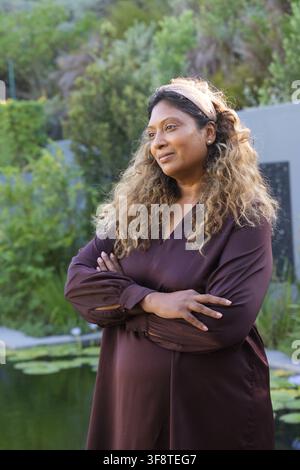 Donna di mezza età in piedi vicino a uno stagno con ninfee in giardino che indossa un vestito bordeaux a maniche lunghe Foto Stock