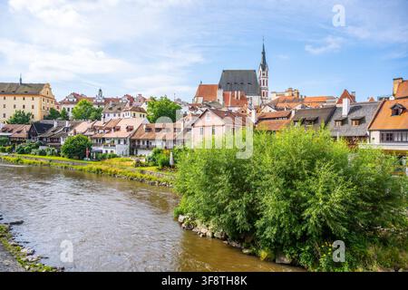 Le persone godono della calda giornata estiva a Český Krumlov. La città vanta edifici colorati e una chiesa sullo sfondo. Il fiume scorre dolcemente accanto alle case storiche. Foto Stock