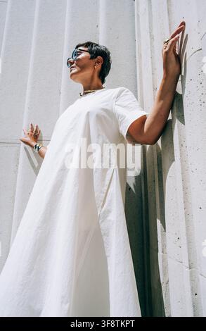 Donna in abito bianco in piedi contro un muro di cemento, evidenziando la moda contemporanea e il design architettonico in un ambiente esterno ben illuminato Foto Stock