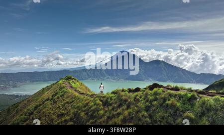 Vista aerea di una persona in piedi su una cresta erbosa che si affaccia sul paesaggio vulcanico del Monte Batur e del Lago Batur a Bali, Indonesia. Foto Stock