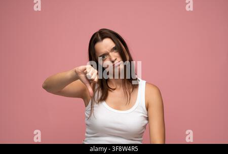 Giovane donna che mostra il gesto del pollice verso il basso per esprimere disapprovazione, rifiuto, antipatia e feedback negativo su sfondo rosa Foto Stock