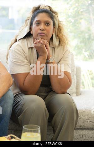 Donna indiana seduta in salotto su un divano chiaro, chinata in avanti con le mani giunte, bicchiere nelle vicinanze Foto Stock