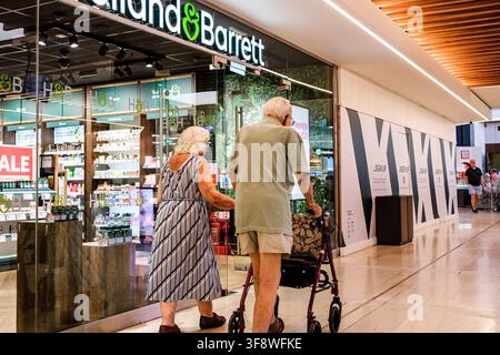 Londra UK, 13 agosto 2026, Vista posteriore di una coppia di anziani che passeggia davanti alla vetrina di un negozio Holland And Barrett, con l’uomo che usa un deambulatore con ruote Foto Stock